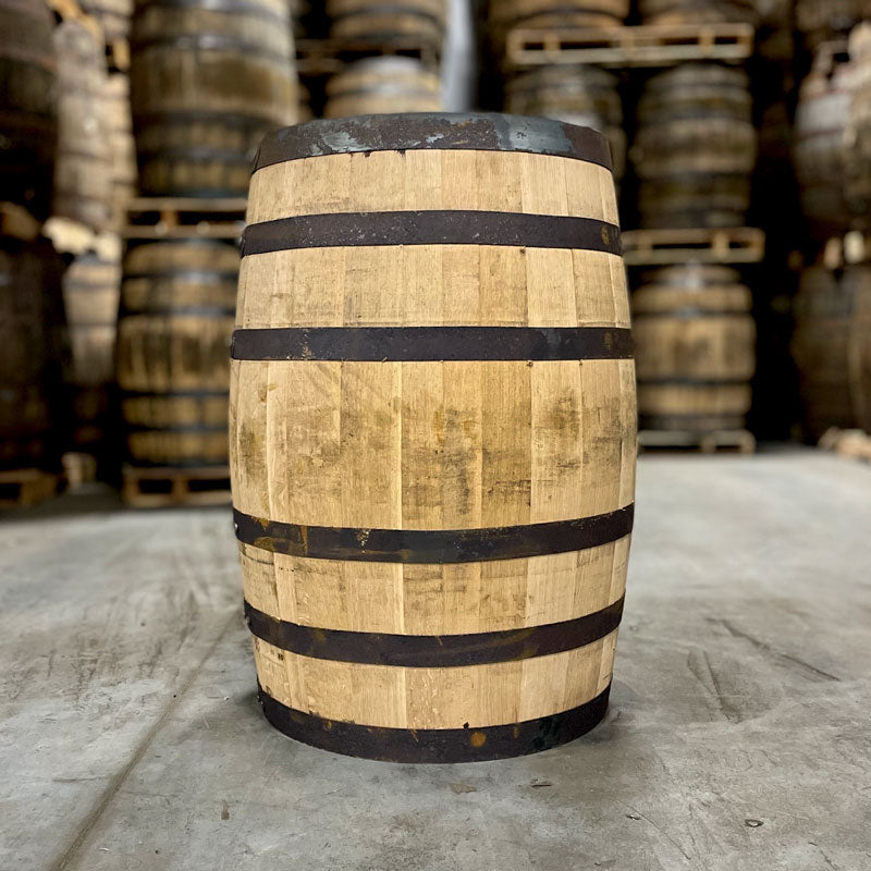 Side of a Penelope Wheated Bourbon barrel with dark rings and other used bourbon barrels stacked on pallets in the background