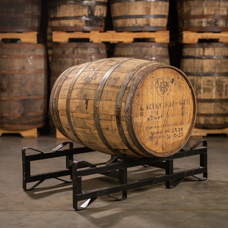 A J Rieger & Co Corn Whiskey Barrel with distillery logo stamped on the head laying on a barrel rack with other used whiskey barrels stacked on pallets in the background