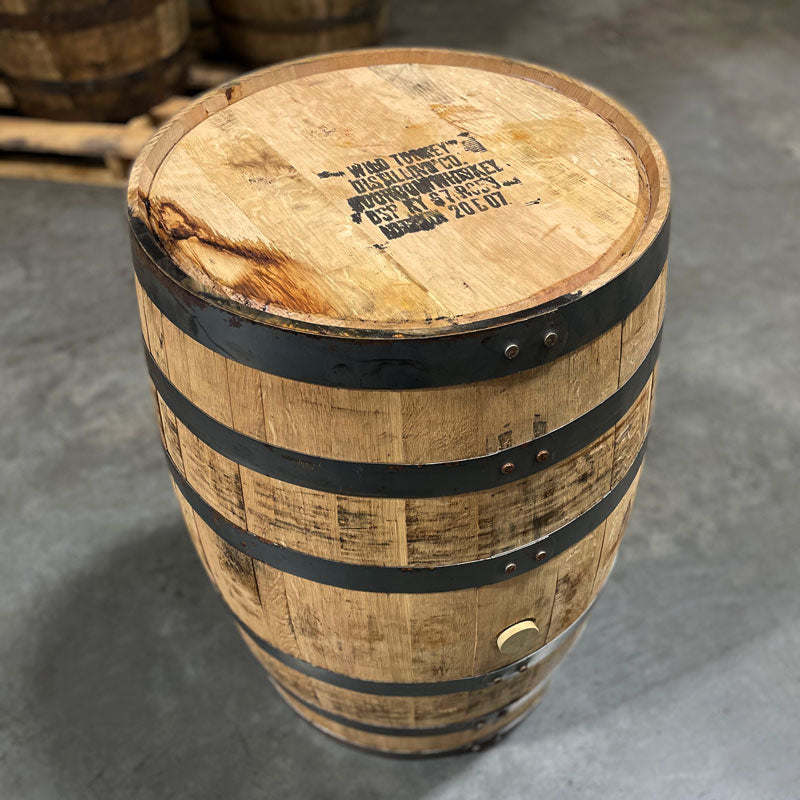 Head and side of a 2-Year Wild Turkey Bourbon Barrel with distillery information and fill date on the head and other used bourbon barrels stacked on pallets in the background.