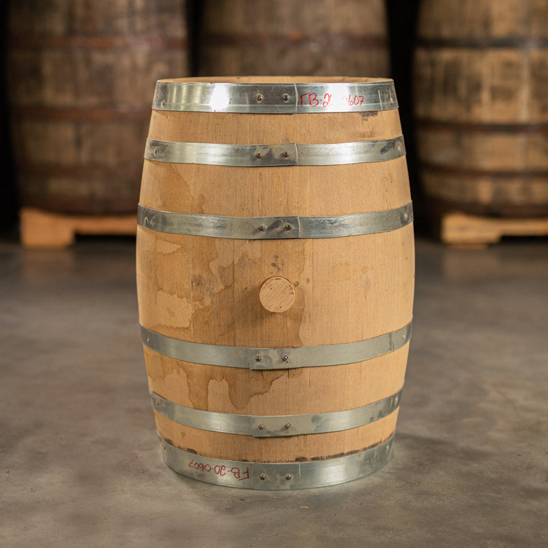 Side of a 15 Gallon Journeyman Bourbon Barrel with shiny, steel rings and other used bourbon barrels stacked on pallets in the background