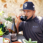 Shannon Harris  taking a sip of beer from a glass while sitting at a table with plants and a bottle of beer