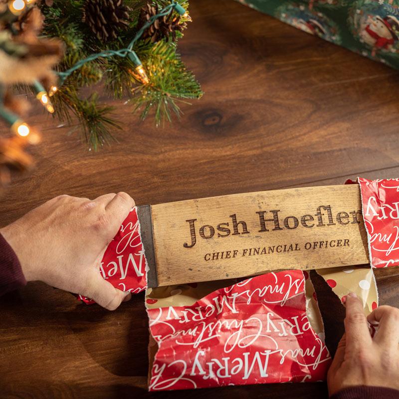 Person unwrapping barrel stave name desk plate under a lit Christmas tree