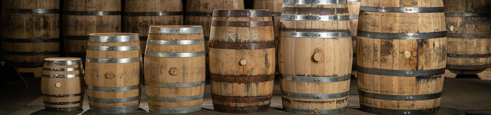 Different sizes of oak aging barrels in front of whiskey barrels stacked on pallets