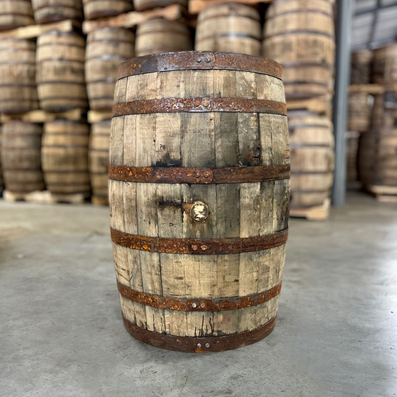 
                  
                    Side of a Booker's Bourbon Barrel with rusted bands and other used bourbon barrels stacked on pallets in the background
                  
                