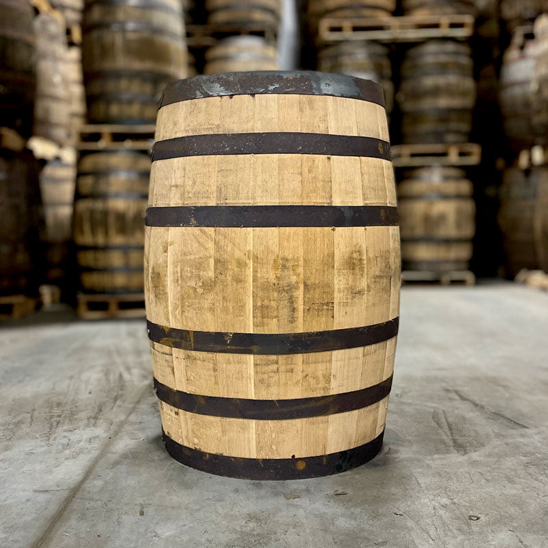 
                  
                    Side of a Penelope Wheated Bourbon barrel with dark rings and other used bourbon barrels stacked on pallets in the background
                  
                