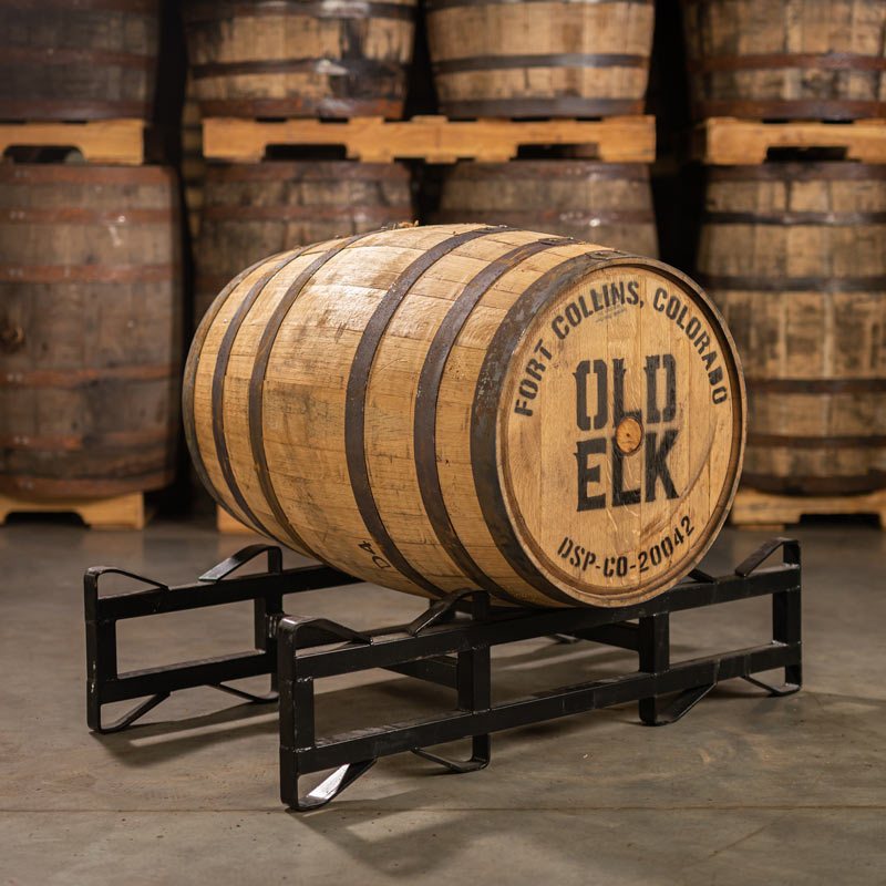 An Old Elk Bourbon barrel with a head bung and distillery stamp laying on a barrel rack with other used bourbon barrels stacked on pallets in the background