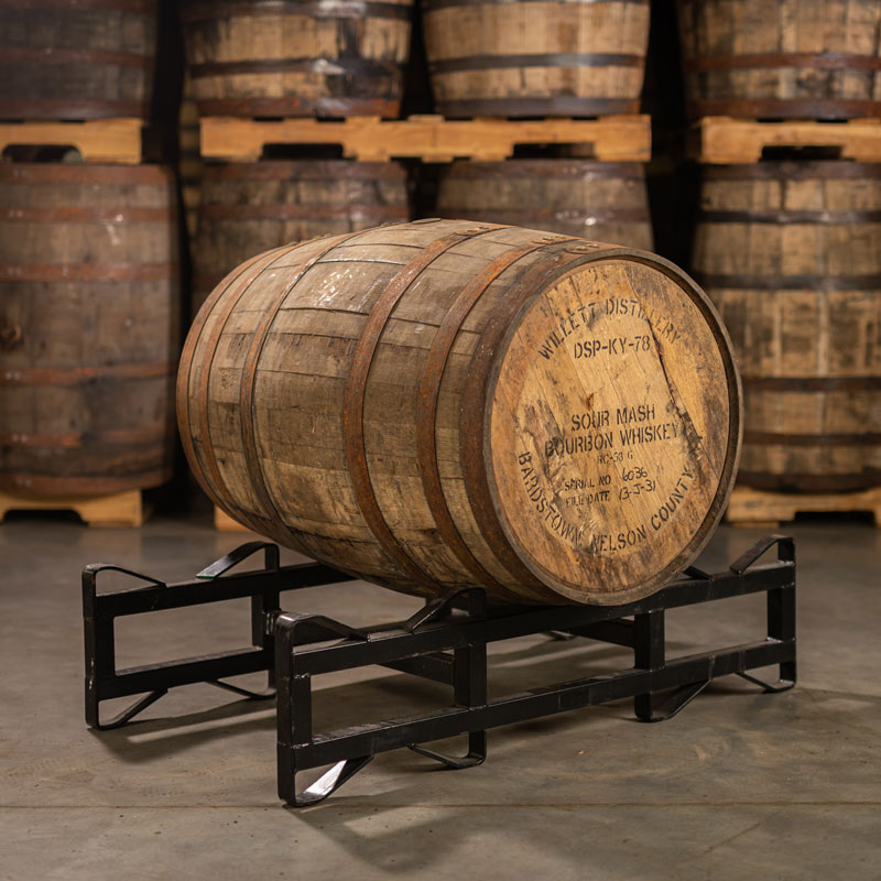 
                  
                    A used 8 year Willett Bourbon barrel on its side on a barrel rack with distillery information stamped on its head and other barrels stacked on pallets in the background
                  
                