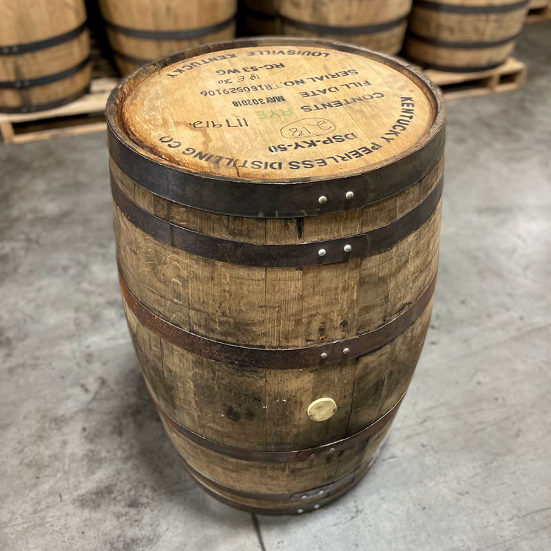 Head and side of a Peerless Rye Whiskey Barrel with distillery name, information and fill date stamped on the head and other used whiskey barrels stacked on pallets in the background.