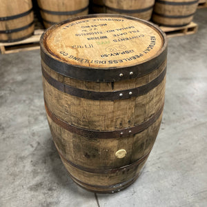 
                  
                    Head and side of a Peerless Rye Whiskey Barrel with distillery name, information and fill date stamped on the head and other used whiskey barrels stacked on pallets in the background.
                  
                