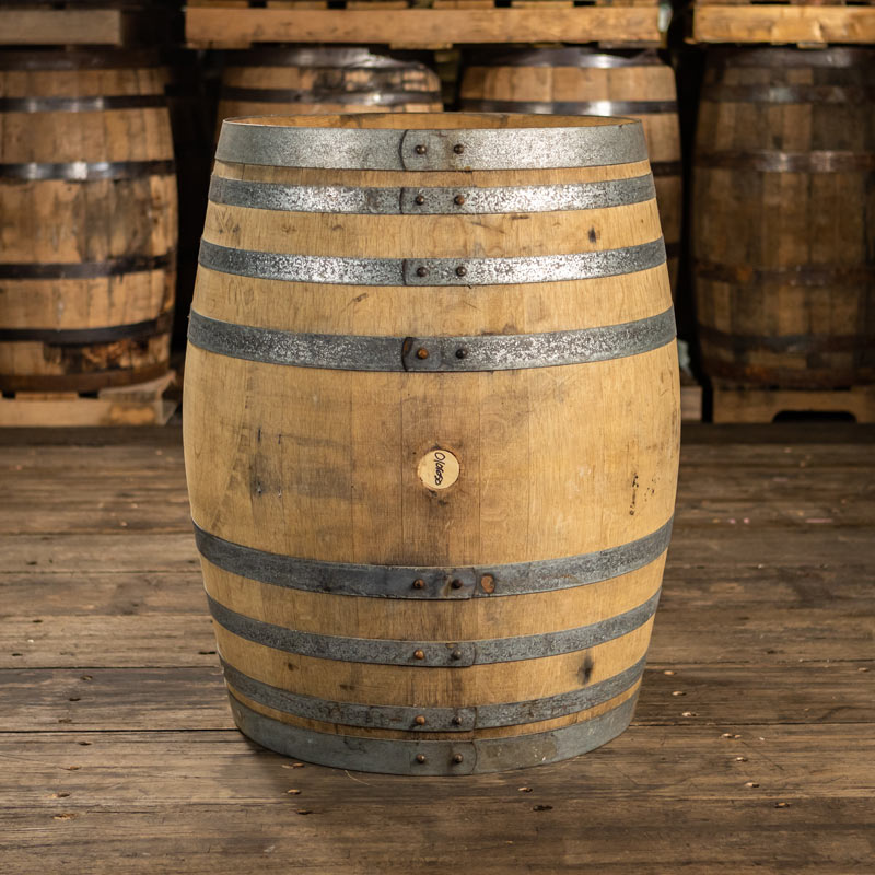 220 liter Oloroso sherry cask with shiny steel rings and light colored staves and barrels stacked on pallets in the background