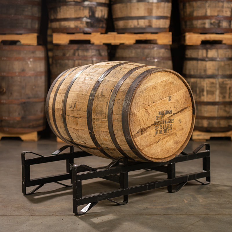 A used 4 year Wild Turkey Bourbon Barrel with distillery information stamped on the head and laying on its side on a barrel rack with other bourbon barrels on pallets in the background