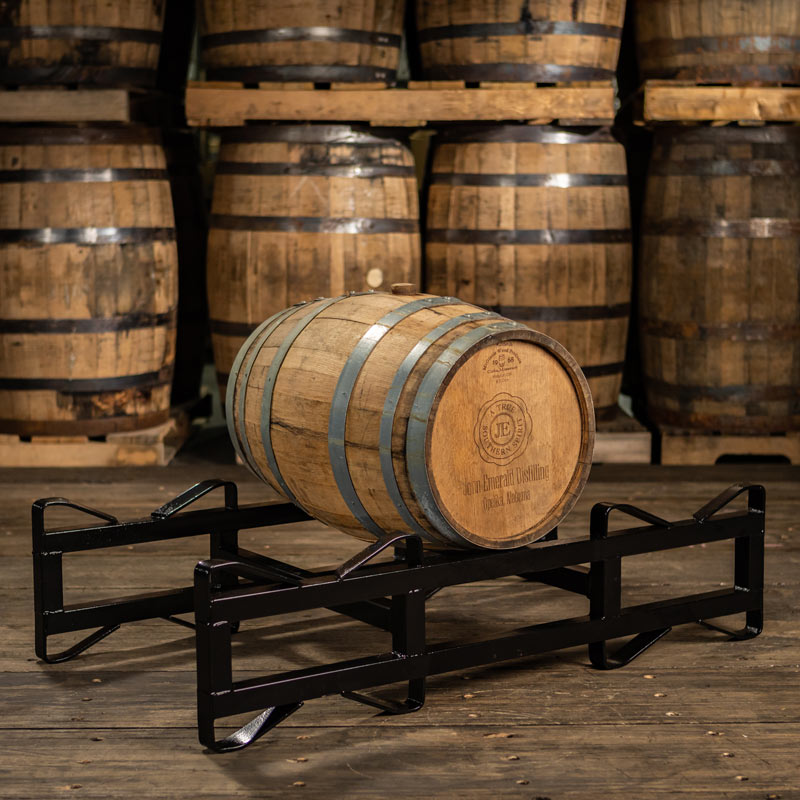 13 Gallon John Emerald Distilling Whiskey barrel on a rack with larger whiskey barrels on pallets in the background