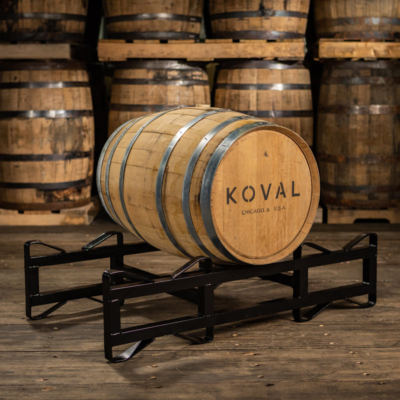 30 Gallon Koval Distillery Rye Whiskey barrel on rack with larger barrels stacked on pallets in the background