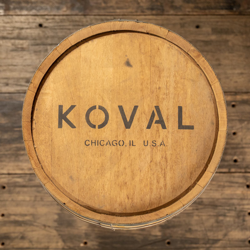 30 Gallon Koval Distillery Rye Whiskey barrel on rack with larger barrels stacked on pallets in the background