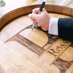
                  
                    The arm of a man in a suit holding a marker in his hand and signing an engraved barrel head wedding guestbook with R initial and Ross family name
                  
                