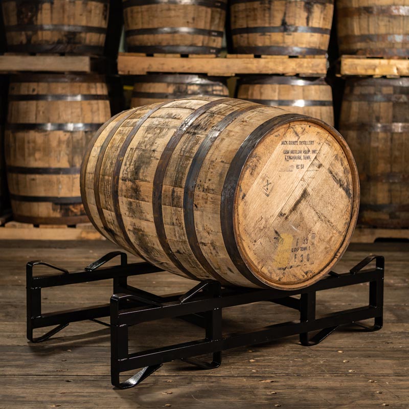 
                  
                    Jack Daniels whiskey barrel on a rack with distillery markings on head, rustic rings and stained staves with barrels stacked on pallets in the background
                  
                